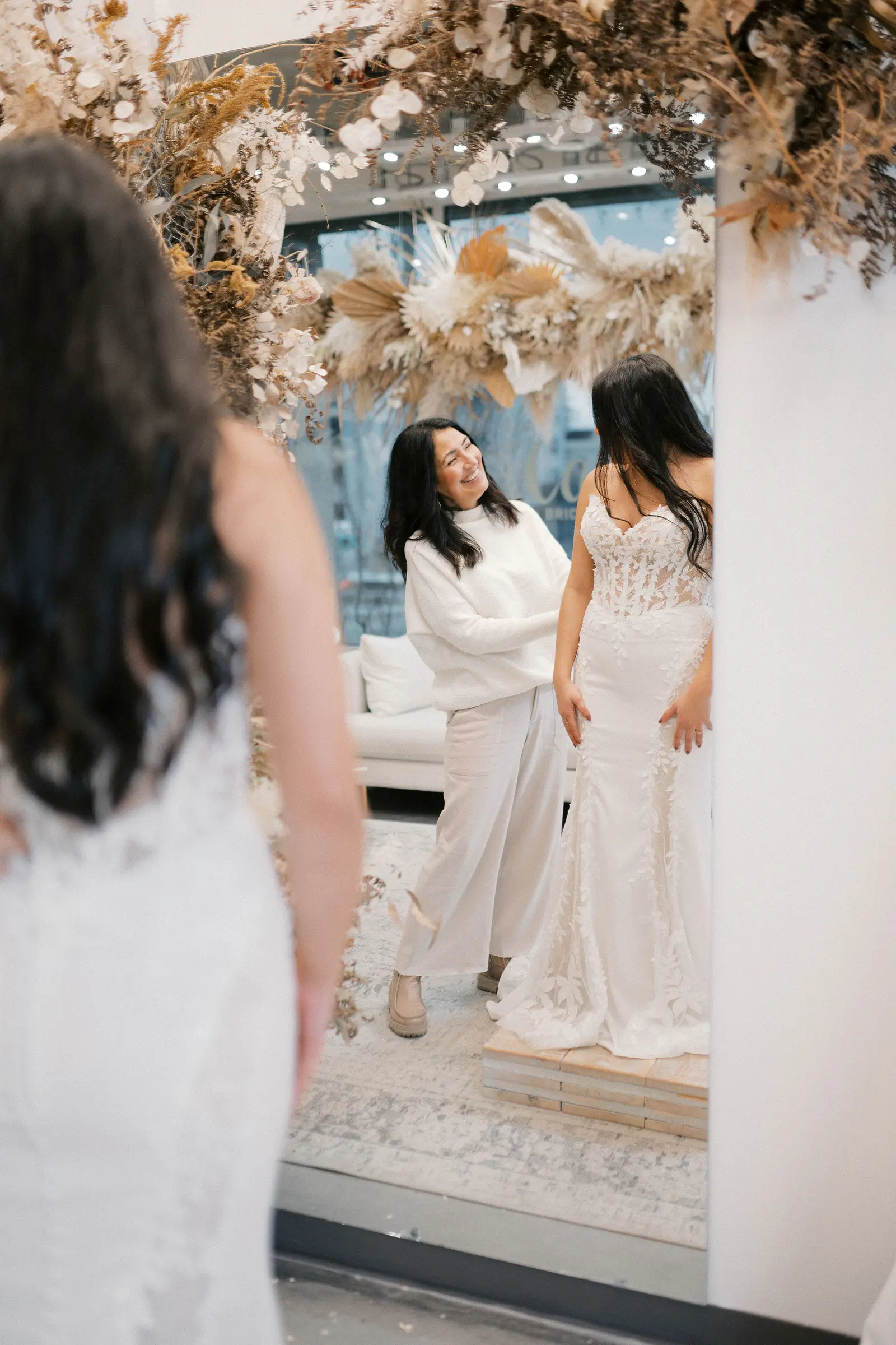 A stylist fastening the back of a gown for a bride during an appointment at a bridal boutique