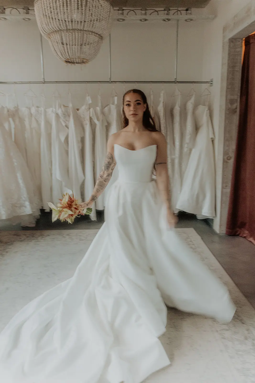 Bride in a strapless Private Collection ballgown with a plain skirt and a long train, standing by the gown rail under a beaded chandelier at an independent bridal boutique