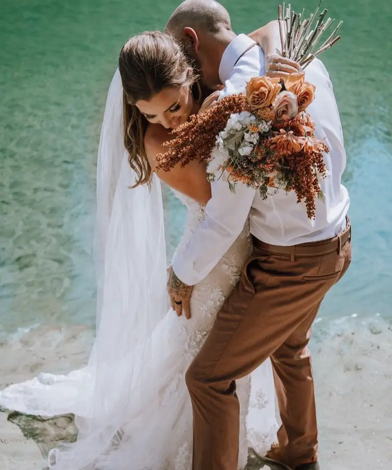 Groom kissing the bride's shoulder at the edge of a turquoise lake, her veil and train across the sand and a rust-coloured bouquet in her hand
